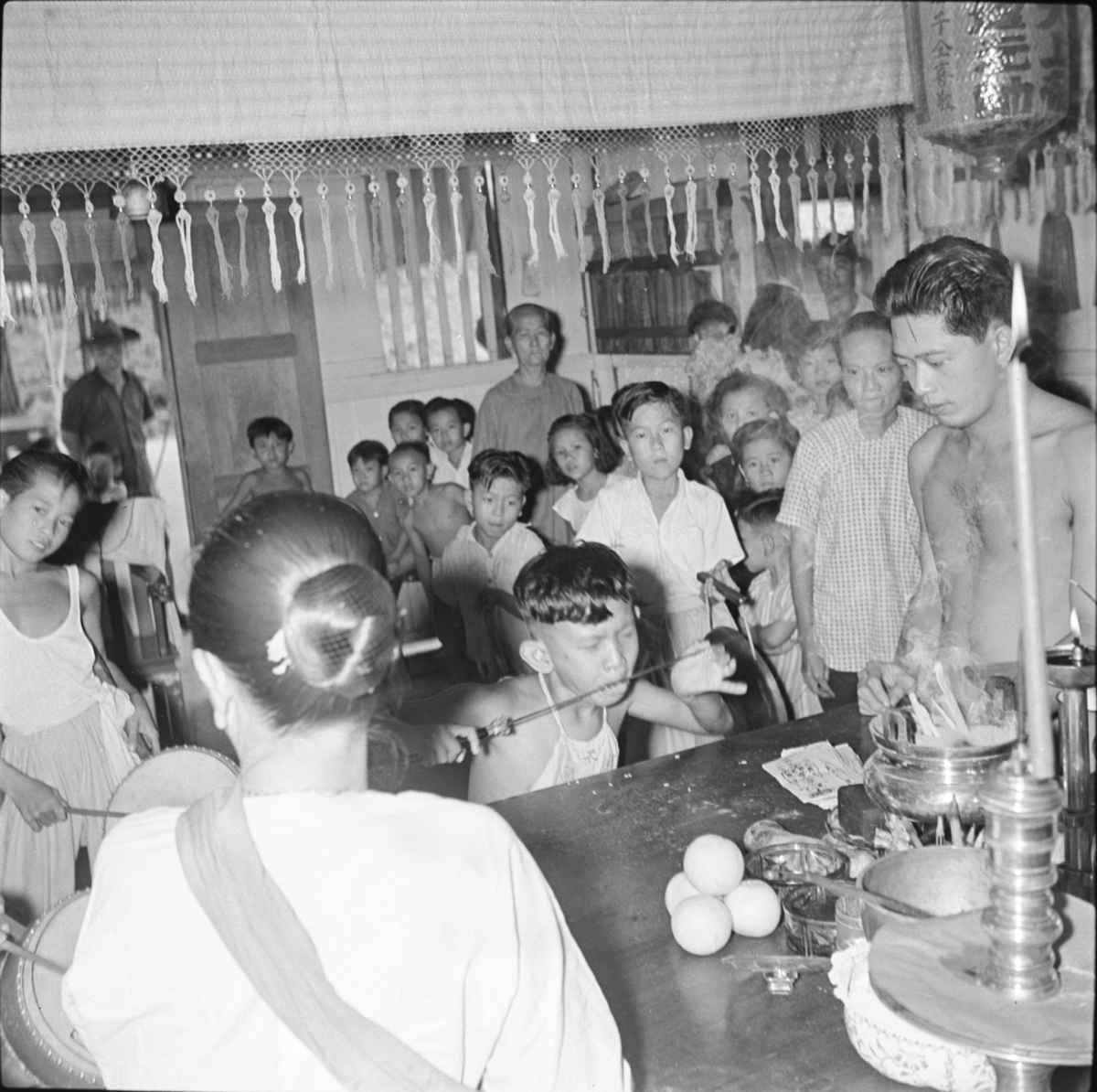 Inside a Chinese temple the spirit-medium, a boy wearing a cloth stomacher stands by an offering table holding a knife to his tongue. Facing him is a woman wearing a blouse and a sash over her left shoulder. A group of men, women and children watch from behind.
