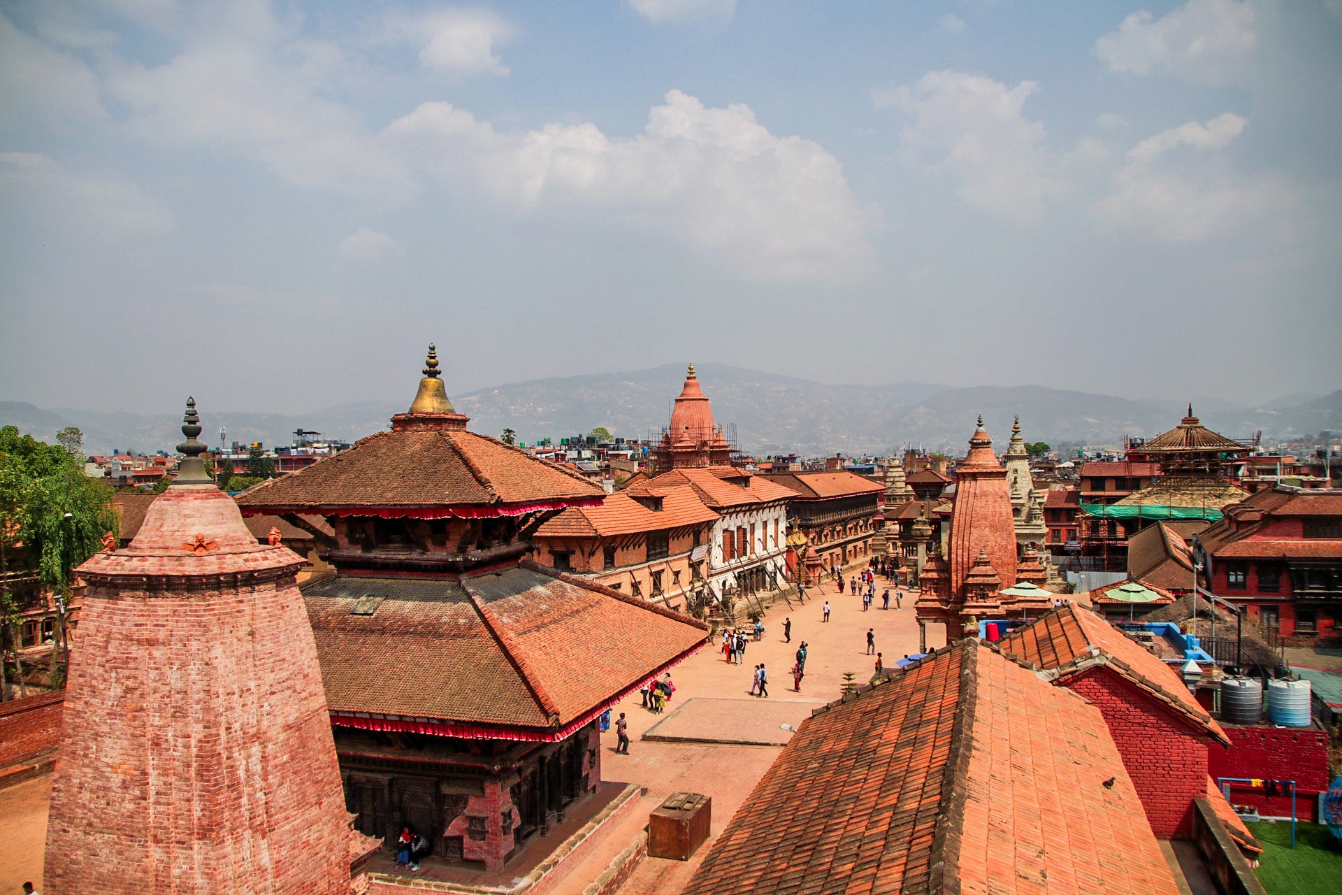This is a photograph of Bhaktapur's skyline taken from the roof of a building. We see multiple temples, all built in brick and wood but the focus in the photograph is on the pagoda and tiered shikhara style roofs of the temples. The city looks visually coherent because of the extensive use of brick to build temples and the uniformity in architectural styles. In the background we see a silhouette of the lower Himalayan mountains and an overcast sky above.