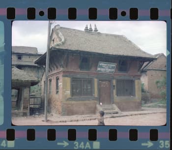 A picture of a building that looks like a one storied house with a roof made in brick. There is a signboard that in Nepali which says it is a women's public library. There are similar looking houses in the background, but they look old and dilapidated.