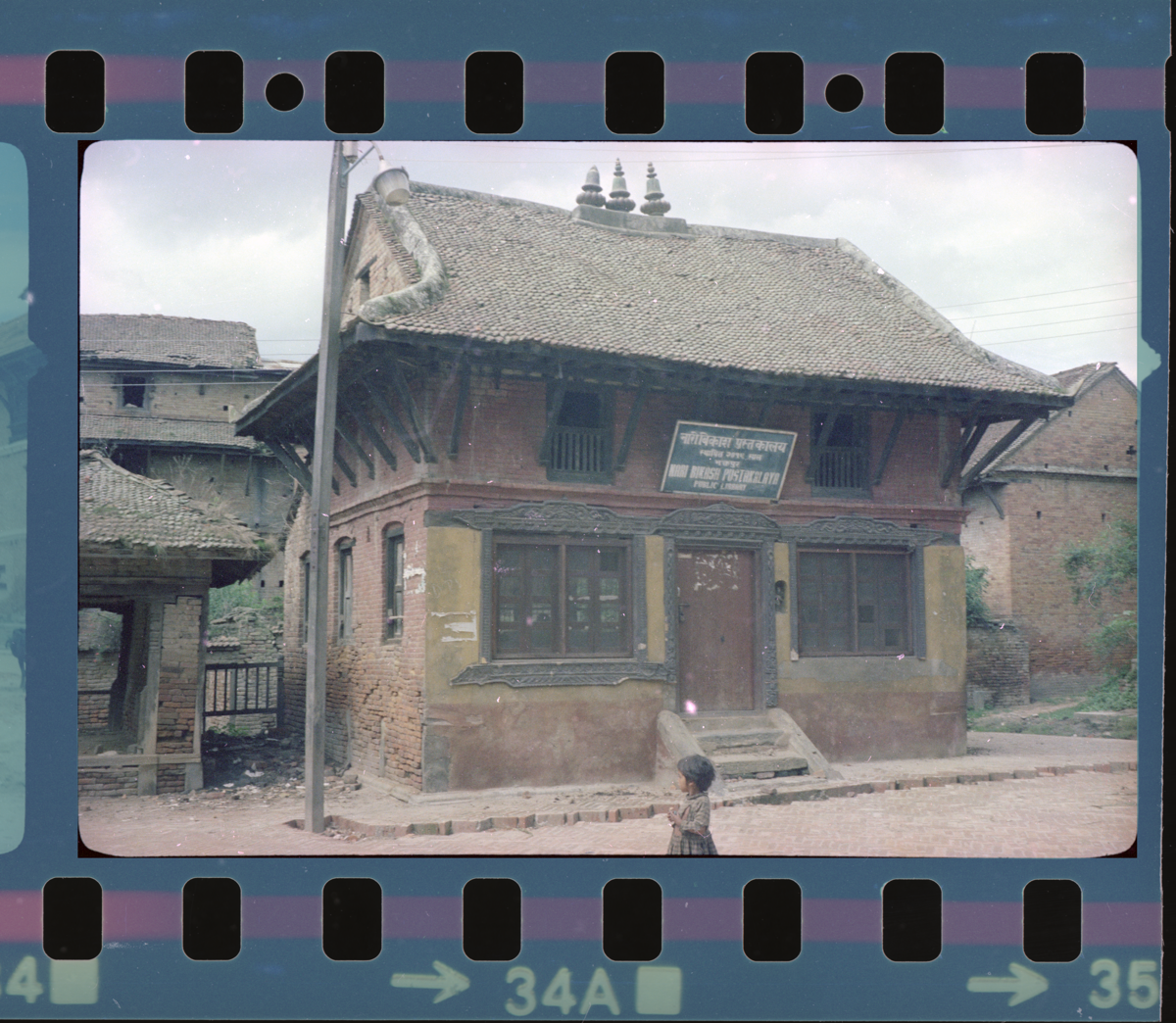 A picture of a building that looks like a one storied house with a roof made in brick. There is a signboard that in Nepali which says it is a women's public library. There are similar looking houses in the background, but they look old and dilapidated.