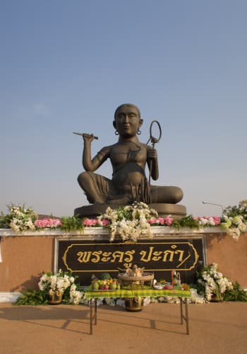 Large, dark grey statue of Pakam. Pakam is a male-presenting figure with a serene expression and dangling earrings. He carries a knife in his right hand and a Pakam rope in his left hand. He is sitting cross legged, with his right knee raised. He is topless, and wearing cloth wrapped around his waist. He is elevated on a platform edged with pink and white flowers. Underneath is a sign that says Phra Khru Pakam in Thai. If on mobile, you will also notice a table under the shrine with various offerings. At the centre is a pig's head.